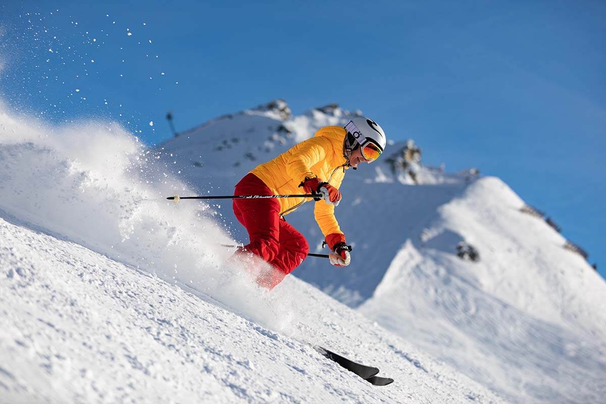 Skiër in oranje jas carvt door verse sneeuw op een zonnige piste
