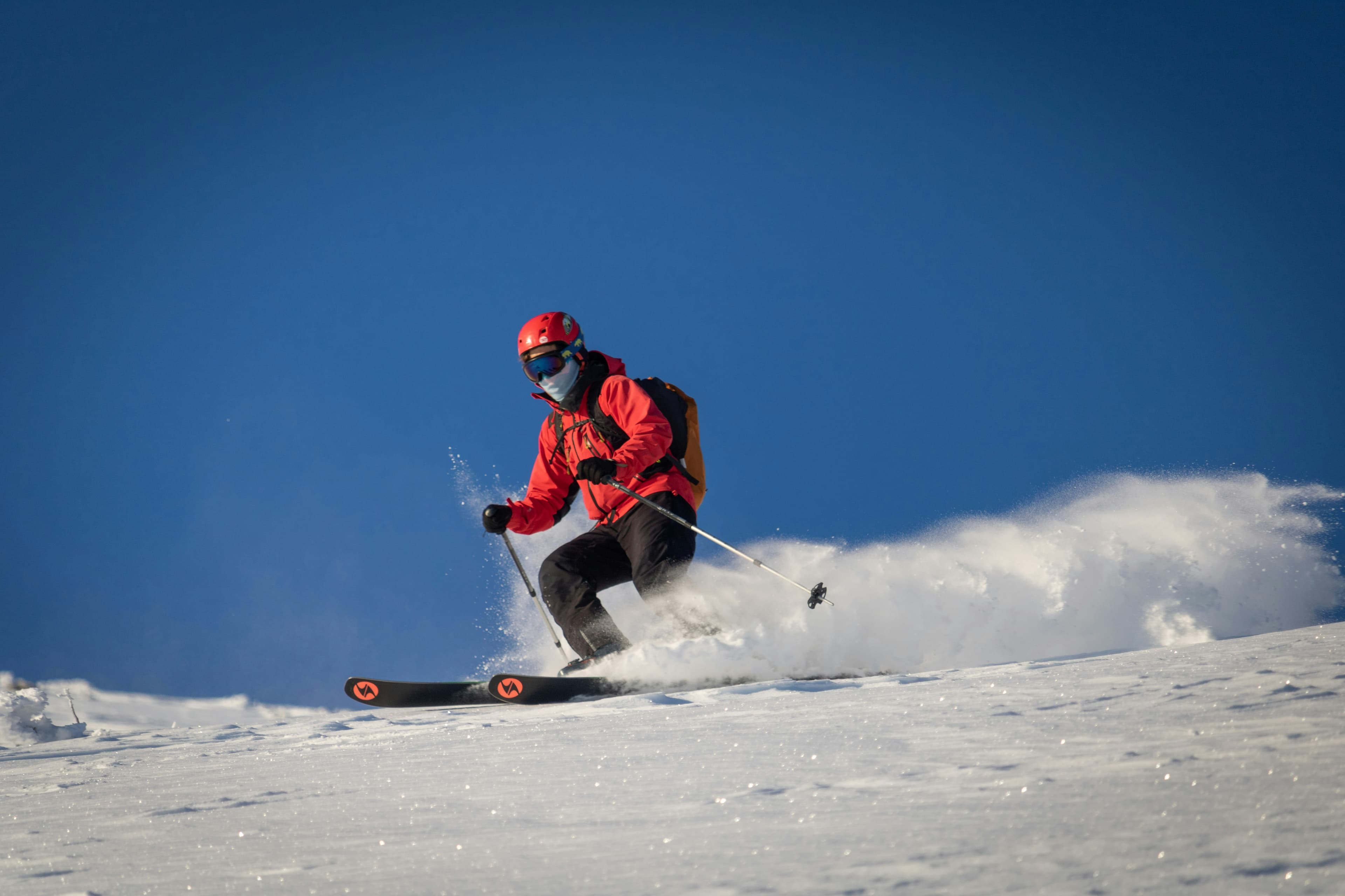 Skiër in rode jas stuift door de sneeuw onder een blauwe lucht